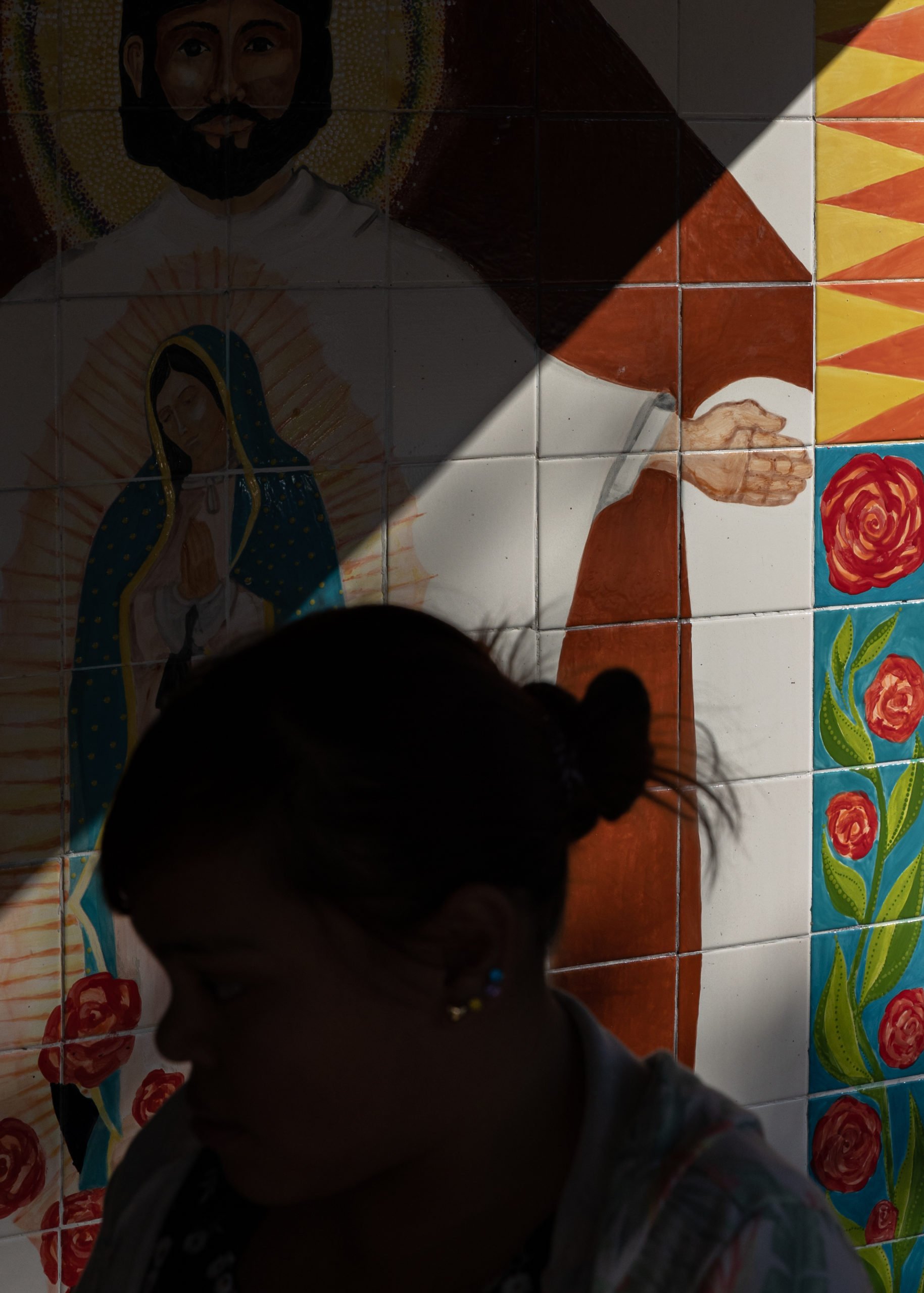 A person's face is silhoutted in shadow in front of a mosaic showing Jesus with outstretched arms, and the Virgin of Guadalupe inside his form. At the edges of the mosiac are painted roses and other bright colored tiles.