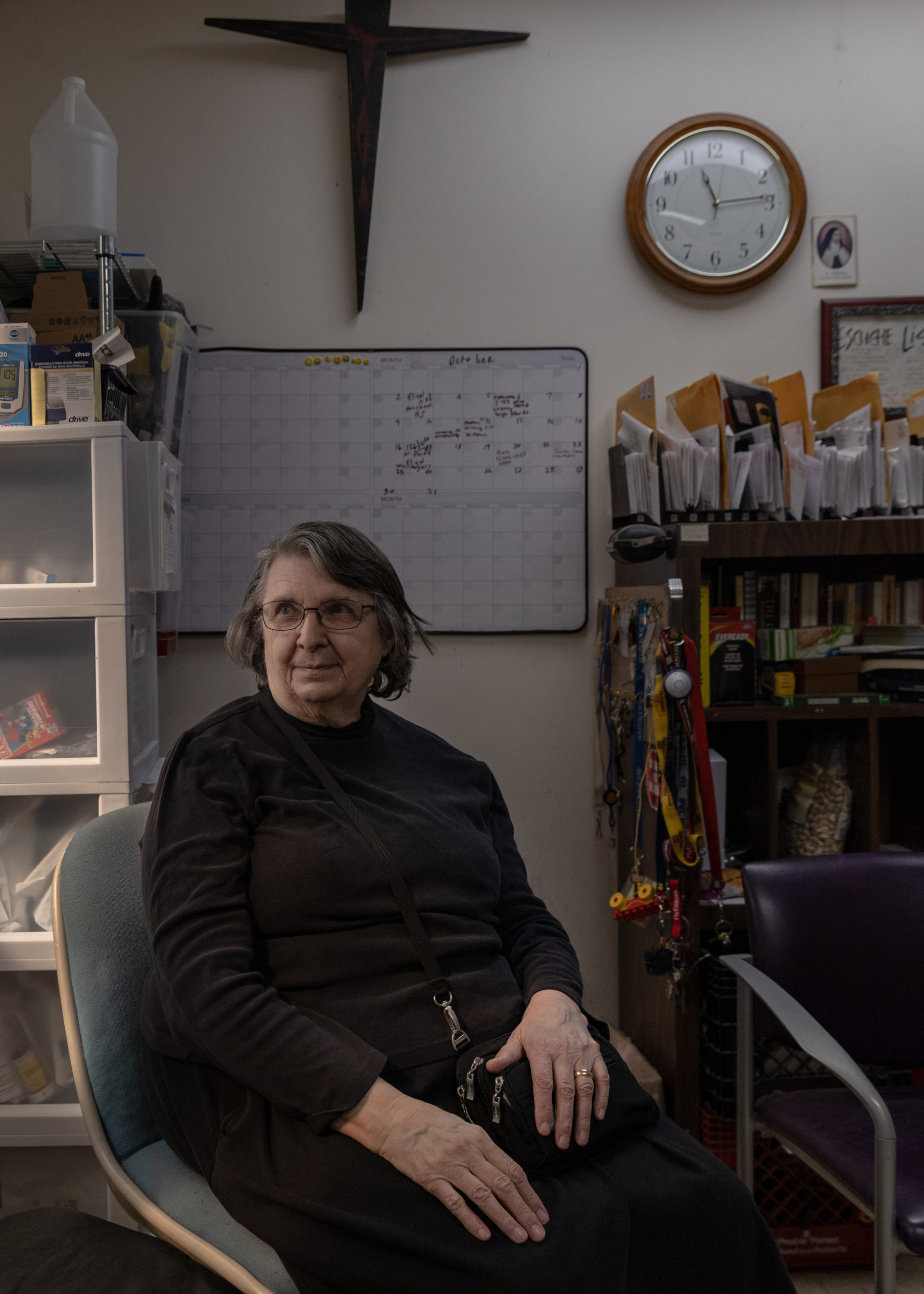 An older woman in dark clothes sits in an office chair among office supplies, a clock, and a prominent but rustic looking wooden cross on the wall.