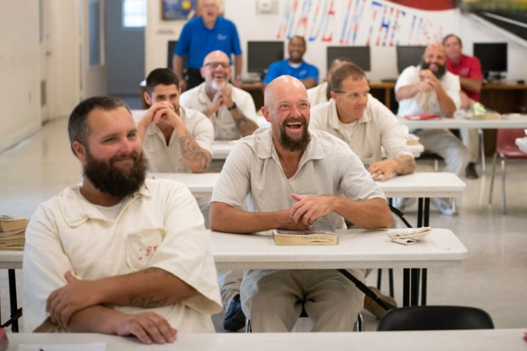 A bald bearded man laughs, seated among other prisoners in a classroom, many of whom are also laughing or smiling. They are all in prison white uniforms, with their instructor visible in a blue shirt behind them.