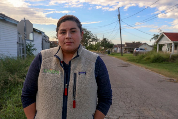 A Latina woman stands, in a light jacket, near worn down houses in a town.
