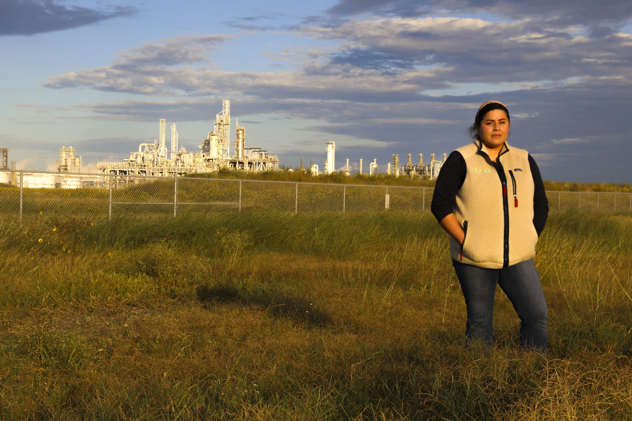 A woman stands with her hands in her pockets, wearing a jacket as she stands in a field of knee-high grass. Behind her in the distance are the industrial buildings of a plastics plant.