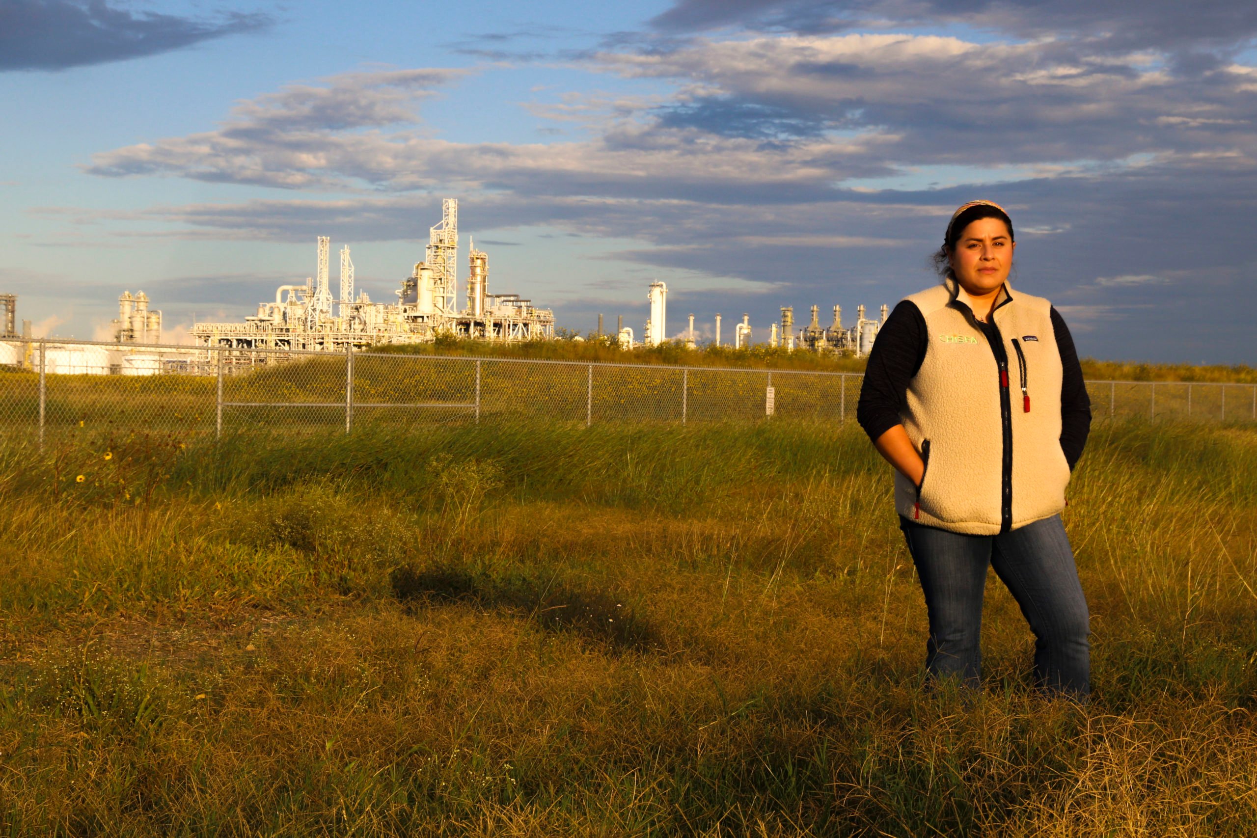 A woman stands with her hands in her pockets, wearing a jacket as she stands in a field of knee-high grass. Behind her in the distance are the industrial buildings of a plastics plant.