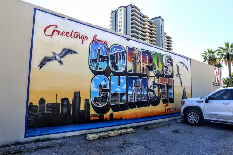A mural reads "Greetings from Corpus Christi" with a seabird in flight and the city skyline and bay depicted.