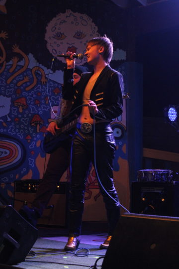 Bowie Brae strikes a dramatic pose with the microphone as he performs with his band Nip Slip at Trans Pride Fest. He's wearing an open jacket with diagonal golden stripes on dark fabric, over a beige crop top, along with a belt with a huge belt buckle over blue jeans. The lighting is dark and moody and another bandmate can be seen behind him.