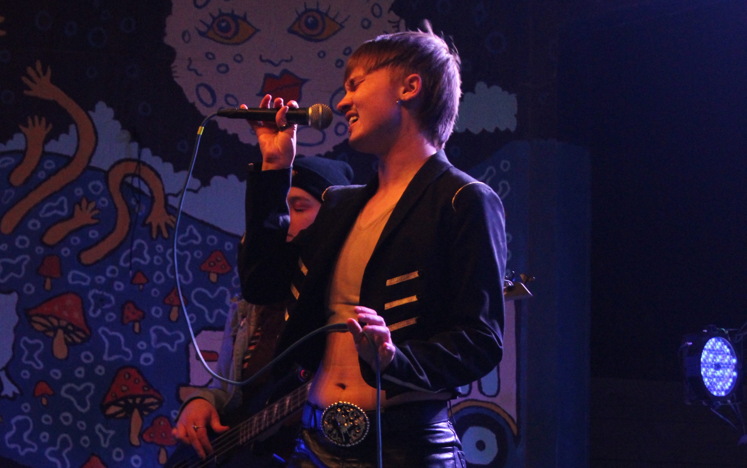 Bowie Brae strikes a dramatic pose with the microphone as he performs with his band Nip Slip at Trans Pride Fest. He's wearing an open jacket with diagonal golden stripes on dark fabric, over a beige crop top, along with a belt with a huge belt buckle.