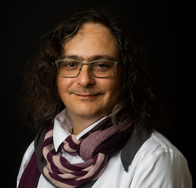 a smiling white person against a dark bg with wavy brown hair, bushy eyebrows, a very light mustache and a big Irish nose and broad forehead. They are wearing a puffy white blouse, dark red and white scarf and green metal wayfarer glasses.