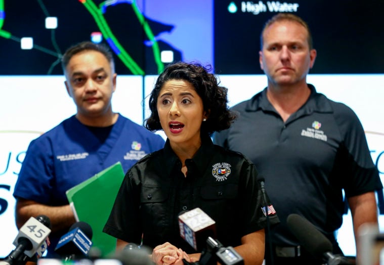 Harris County Judge Lina Hidalgo, wearing a black uniform polo shirt, is flanked by two other officials as she speaks into a bank of microphones.