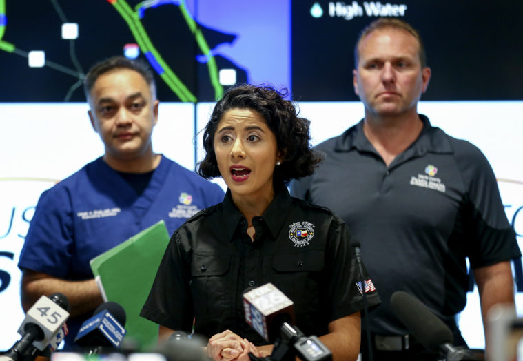 Harris County Judge Lina Hidalgo, wearing a black uniform polo shirt, is flanked by two other officials as she speaks into a bank of microphones.