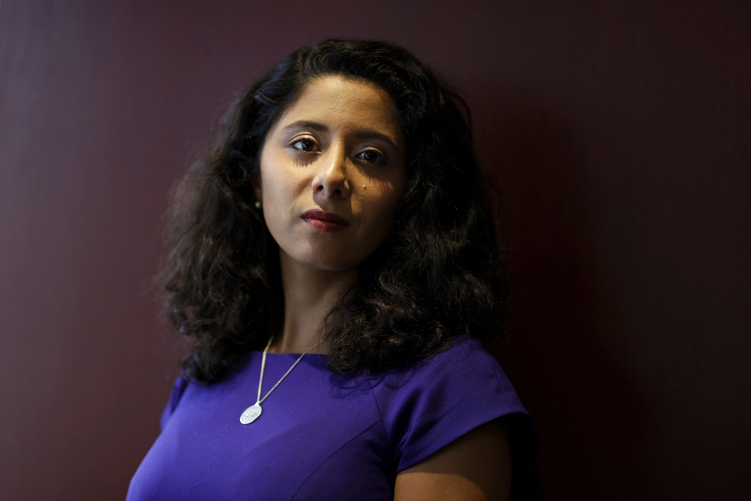 Lina Hidalgo, a Hispanic woman in a blue top with a small round pendant necklace, poses for a portrait, her shoulder length dark hair framing her face.