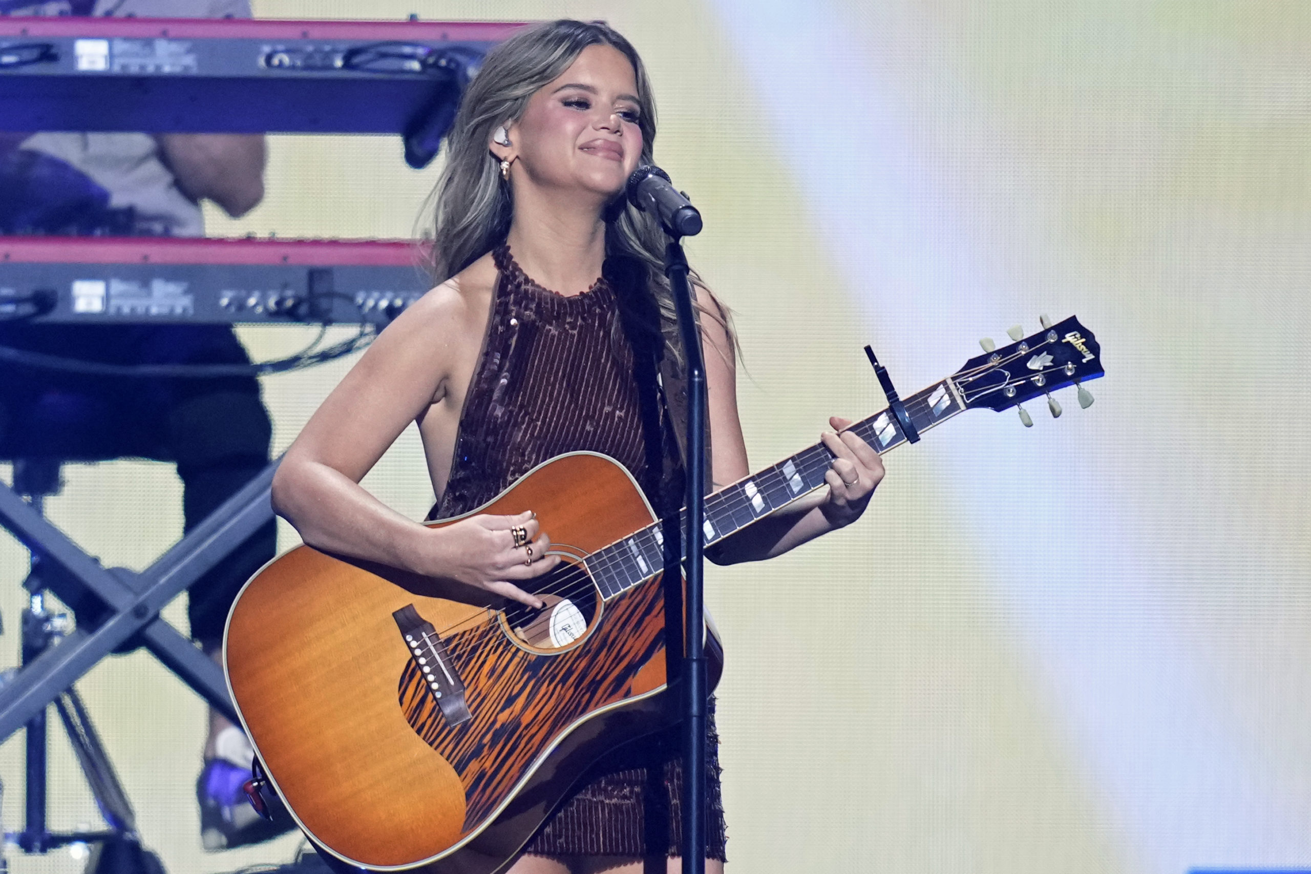 Maren Morris smiles as she sings and plays a guitar at a micrphone. She's wearing a sleeveless black dress.