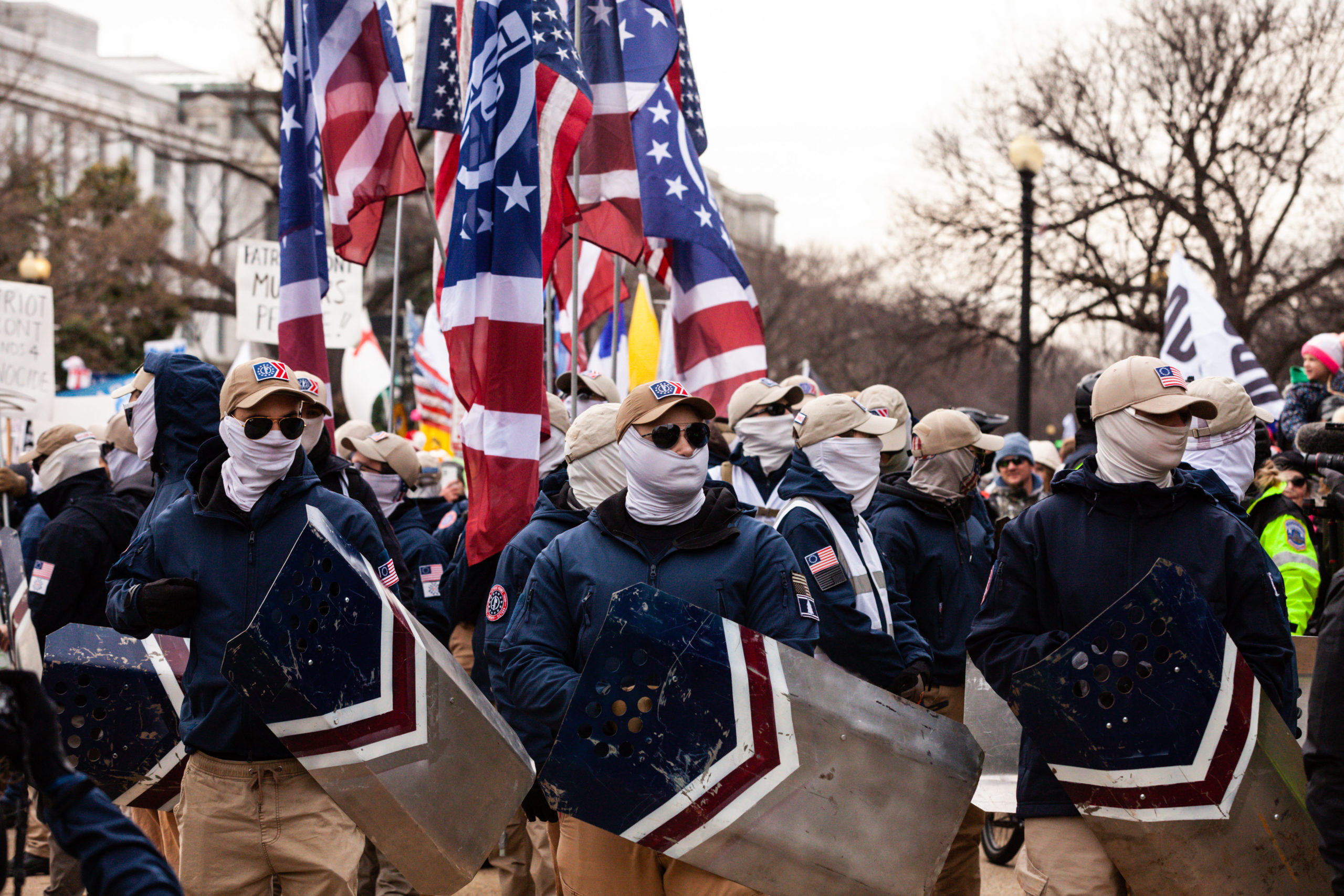 Patriot Front members stand in formation as they prepare to march, wearing blue jackets and white gaiters. They are carrying blue and white shields, and modifed American flags with a fasces on them. Patriot Front now face a lawsuit relating to vandalism of a mural.