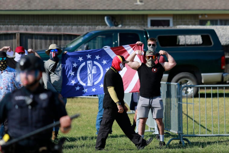 Patriot Front members posture in their uniforms of polo shirt and gaiter, with one holding up his muscular arms in a threatening yet cartoonish pose. Their flag is red, white and blue with a fasces prominently featured at its center.