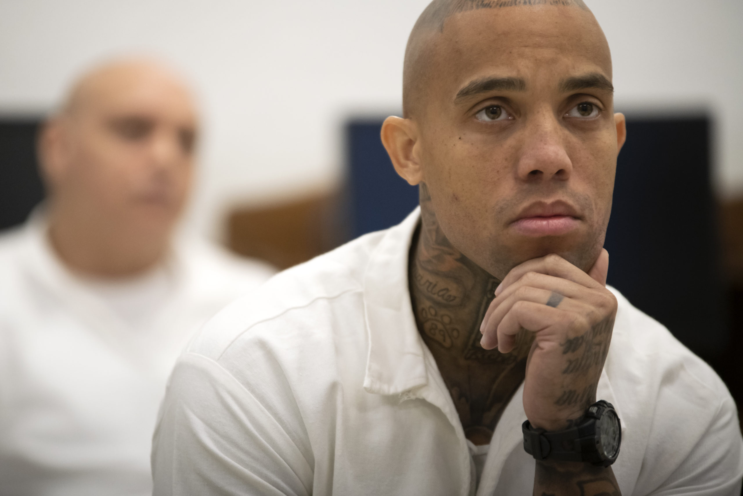 A bald, tattooed BIPOC man sits with his chin in hand, following along with a prison education class with rapt attention. He's wearing a watch and a white prison uniform shirt, and another prisoner is visible behind him.