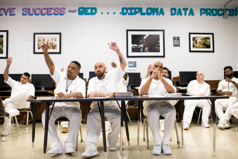 Three inmates have their hands raised, seated in a classroom with about 6 total students visible. Motivational words (Success, GED, Diploma, etc) and photos of other classes decorate the back wall.