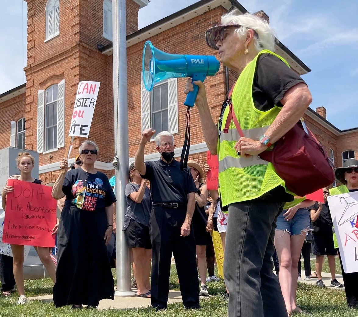 Rev. Carter Heyward, an older white woman in dark pants and a yellow safety vest, speaks into a microphone. In the background is a proterst or rally.