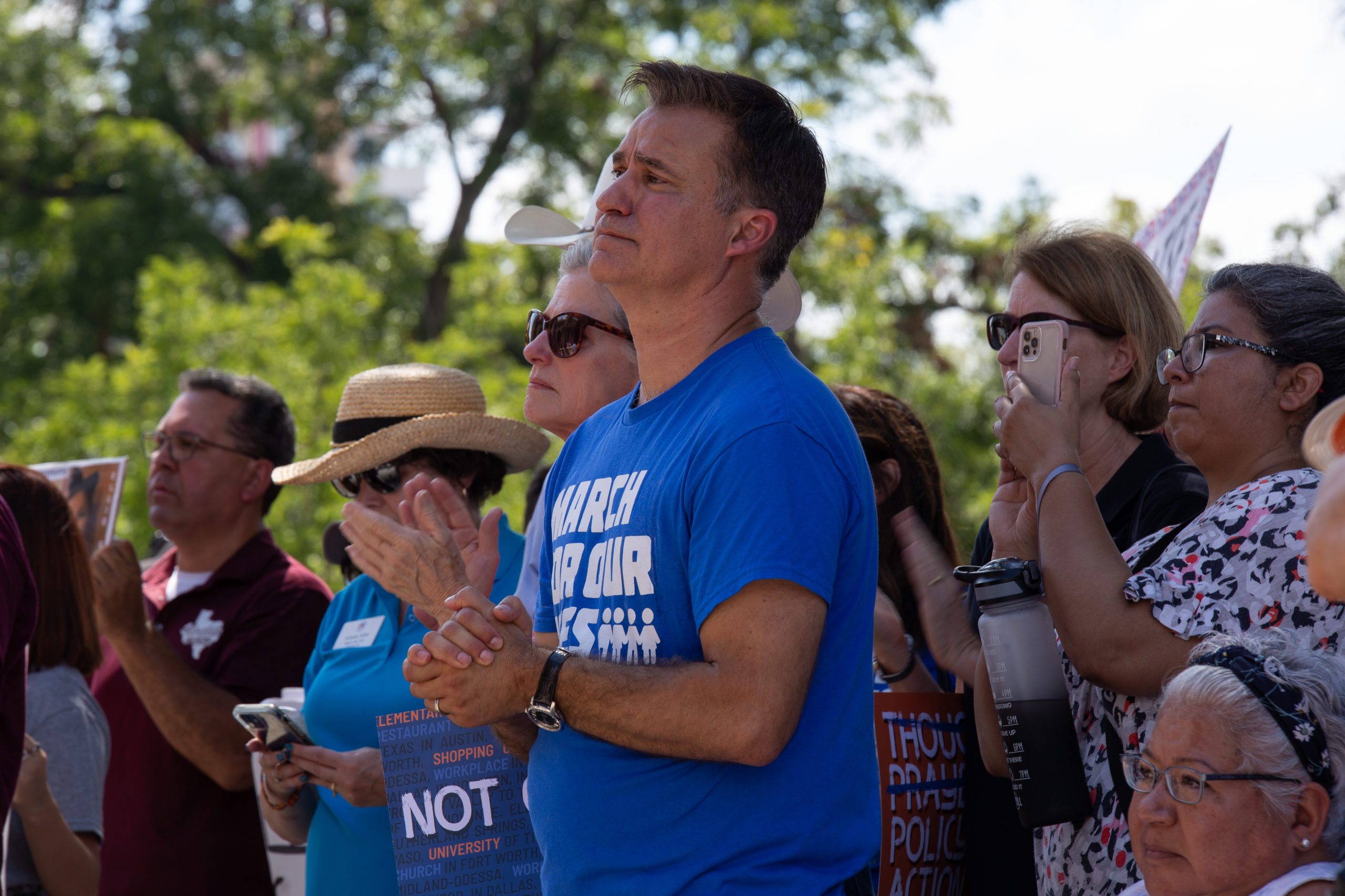 Roland Gutierrez stands with hands clasped, wearing a blue March For Our Lives shirt. He's surrounded by parents and supporters of the Uvalde families at a rally at the Texas Capitol.