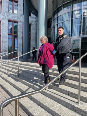 After the oil export sit-in at the U.S. Department of Transportation, a police officer escorts an older woman with white hair, in a red jacket and handcuffs, down the stairs of a municipal buiding.