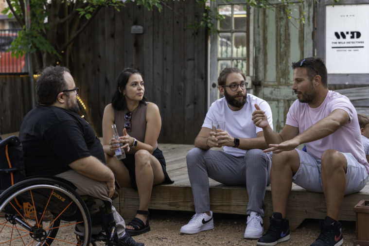 Fou people sit on the edge of a wooden boardwalk style walkway in the backyard of the Wild Detectives in Dallas, Texas