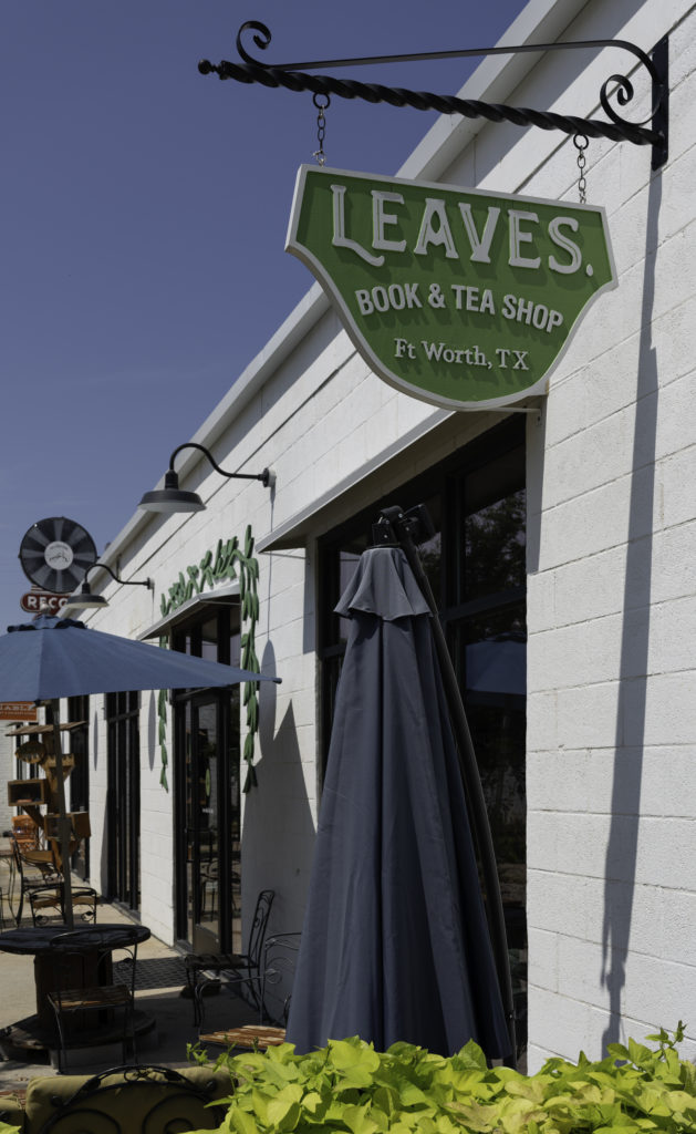 Patio seating with umbrellas outside the Leaves Book & Tea Shop, which has its green sign hanging in the foreground of the photo. There are potted plants visible and even little leaf decorations around the doorway.