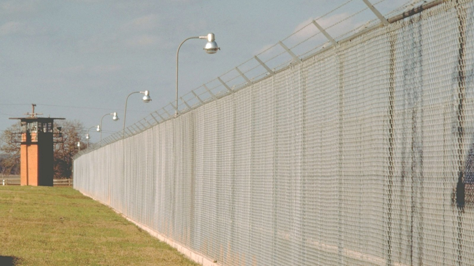 A tall prison wall with barbed wire at the top, lights at regular intervals and a guard tower.