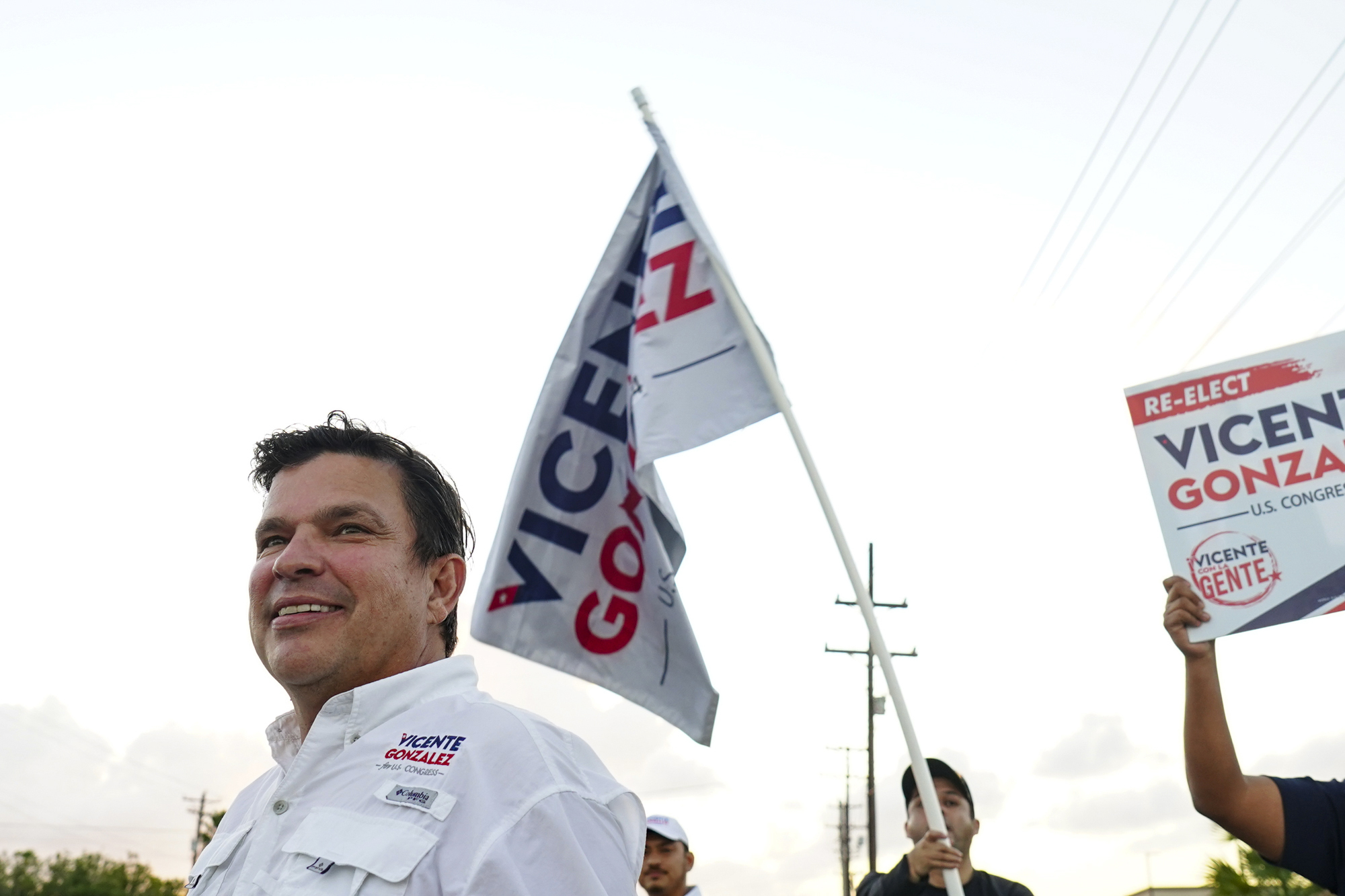 Vicente Gonzalez, a Latino man with short brown hair, smiles while wearing a campaign-branded button down shirt, a Gonzalez flag and sign held aloft behind him.