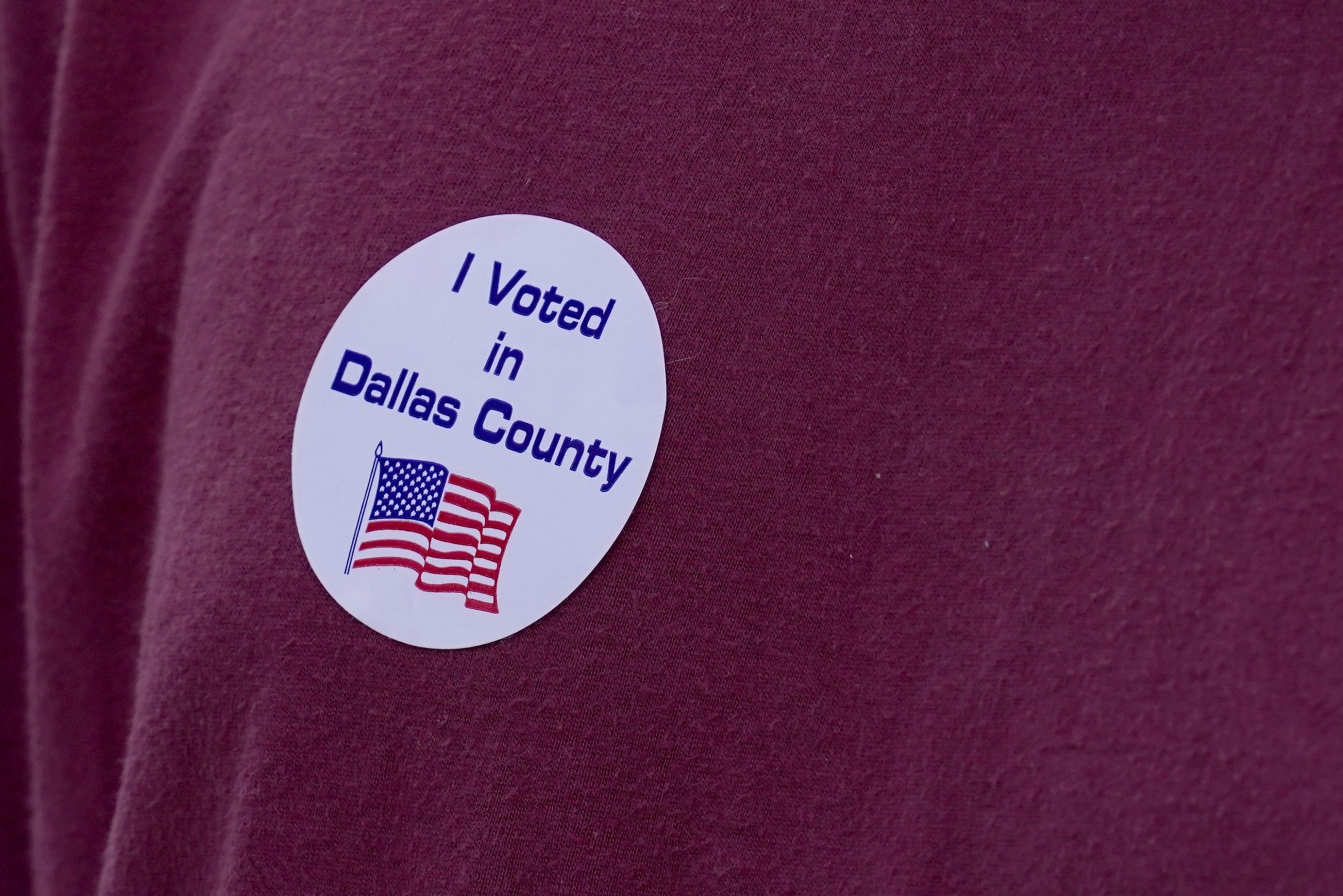 A closeup of a person's red shirt with a sticker: I voted in Dallas County. The sticker also has a wavy US flag.