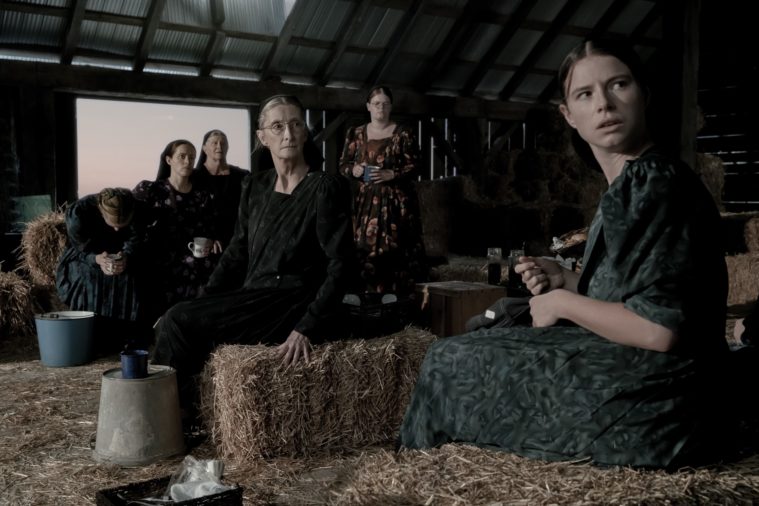 The cast of Women Talking, dressed in simple Mennonite clothing, sit in a barn, looking towards and to the right of the camera.