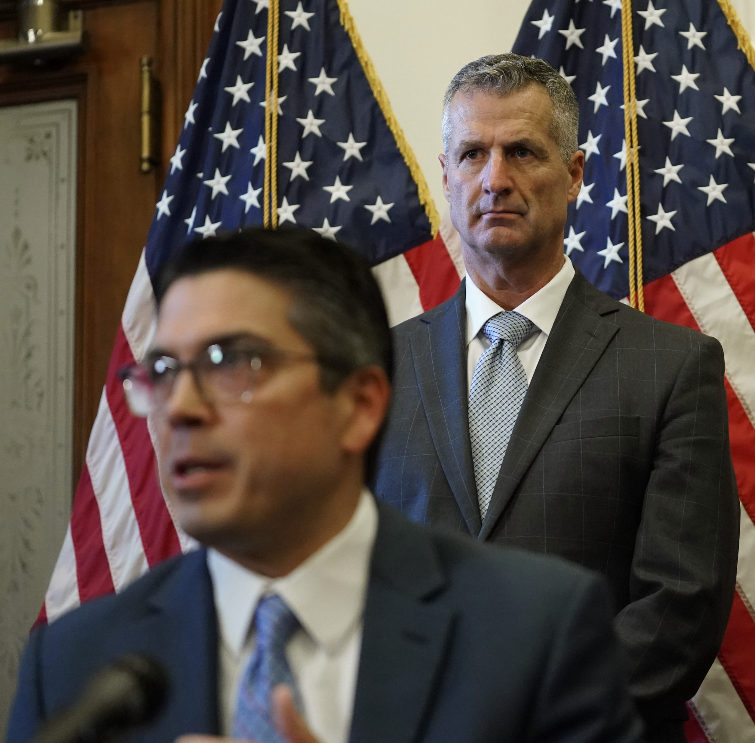 State Representative Chris Paddie speaks into a microphone as ERCOT President and CEO Brad Jones stands behind him, just in front of some United States flags.