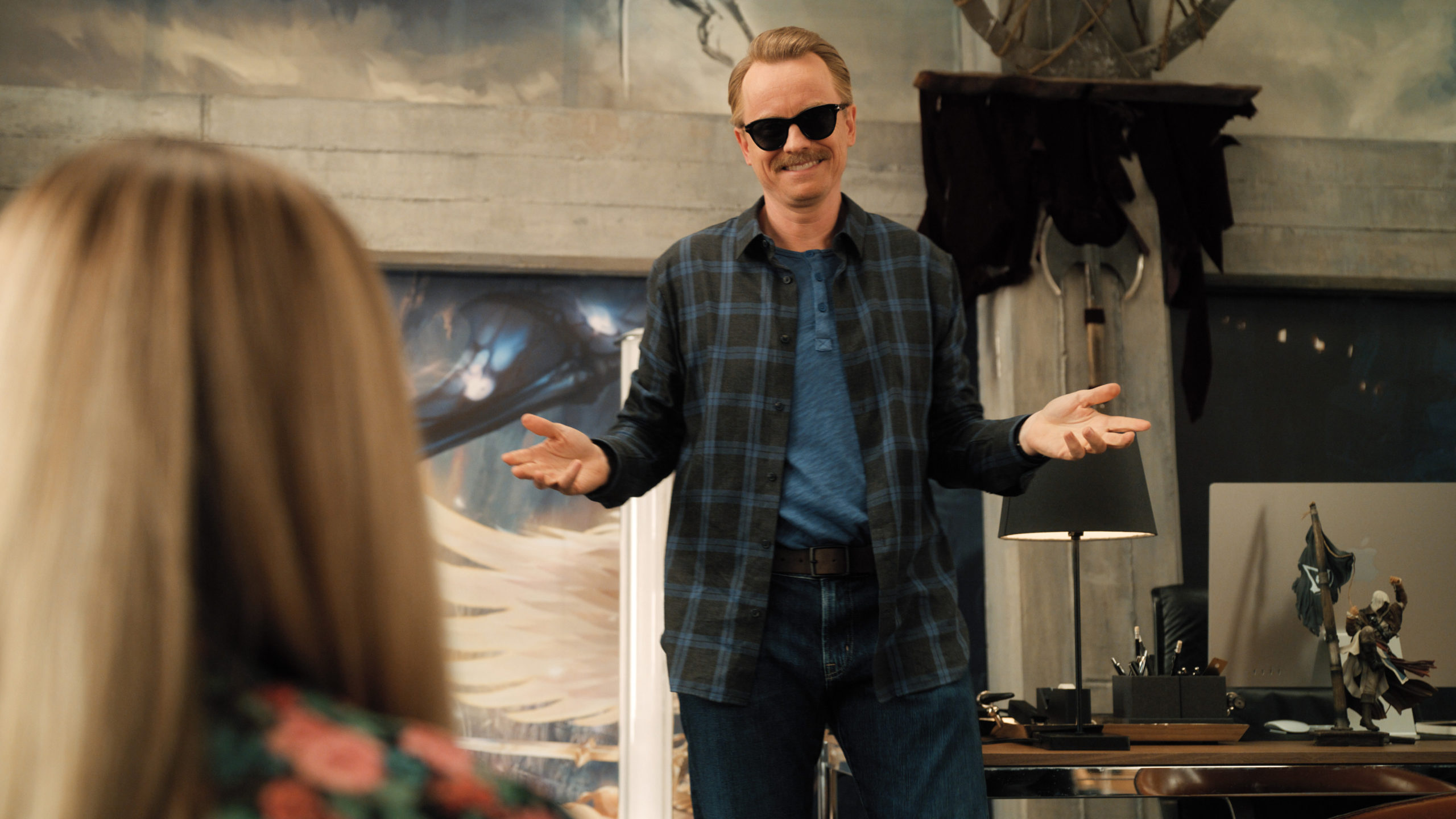 David Hornsby, a white guy with a mustache, holds his hands out at his sides, palms up. He's wearing a long dark blue flannel shirt over dark pants, and sunglasses.