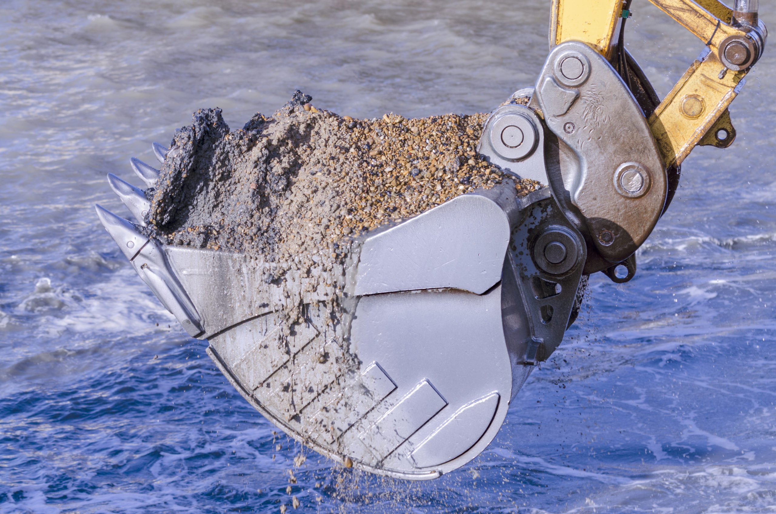 The bucket of an excavator dredges up mud and gravel at the edge of the water.