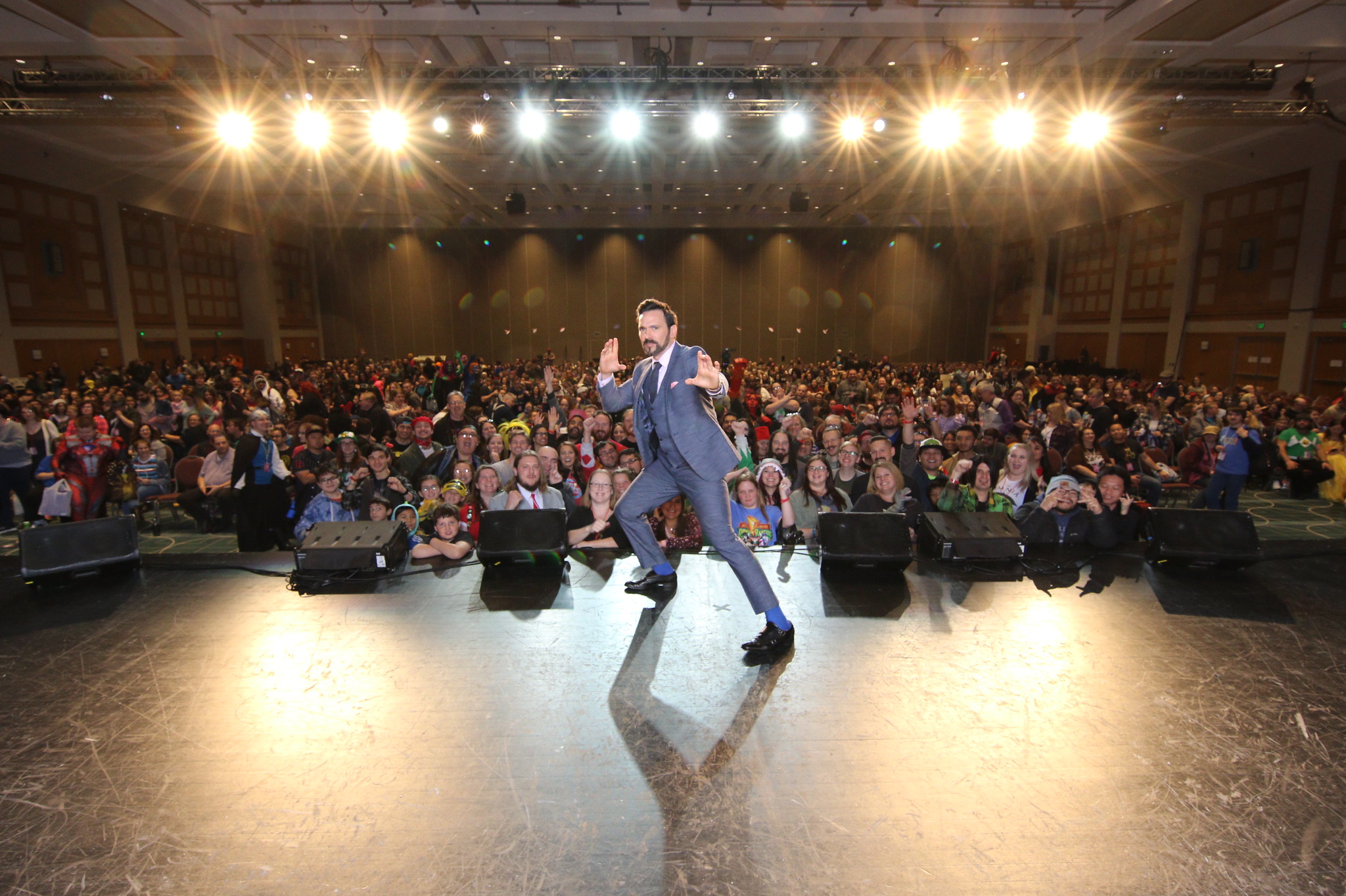 Dressed in a suit, Jason David Frank strikes a dramatic pose on stage, hands held in martial-arts chopping poses, as a crowd looks on with excitement behind him, smiling for the camera.