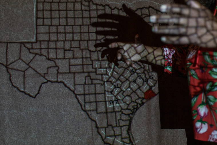 The outline of Texas appears on a cracked, dry piece of fabric which looks like drought-ridden earth. Jones' hands cast shadows on the wall in the projector's light.