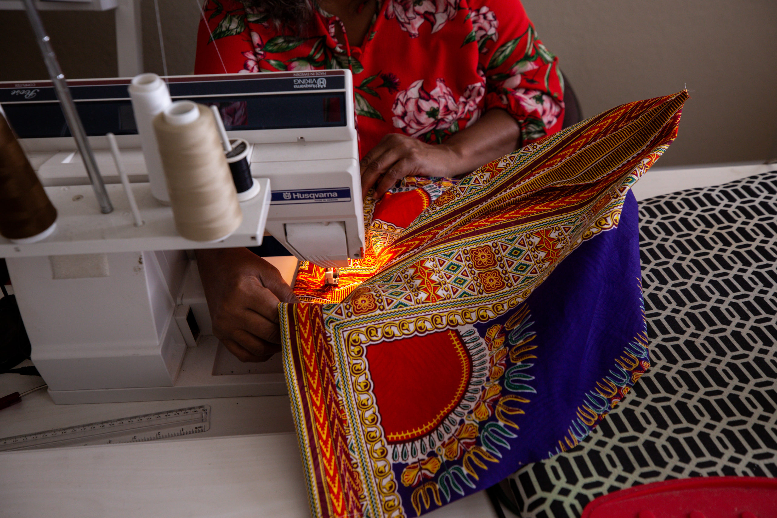 A Black woman, seen as her torso and hands working on a sewing machine, with colorful red and gold fabric being fed through.