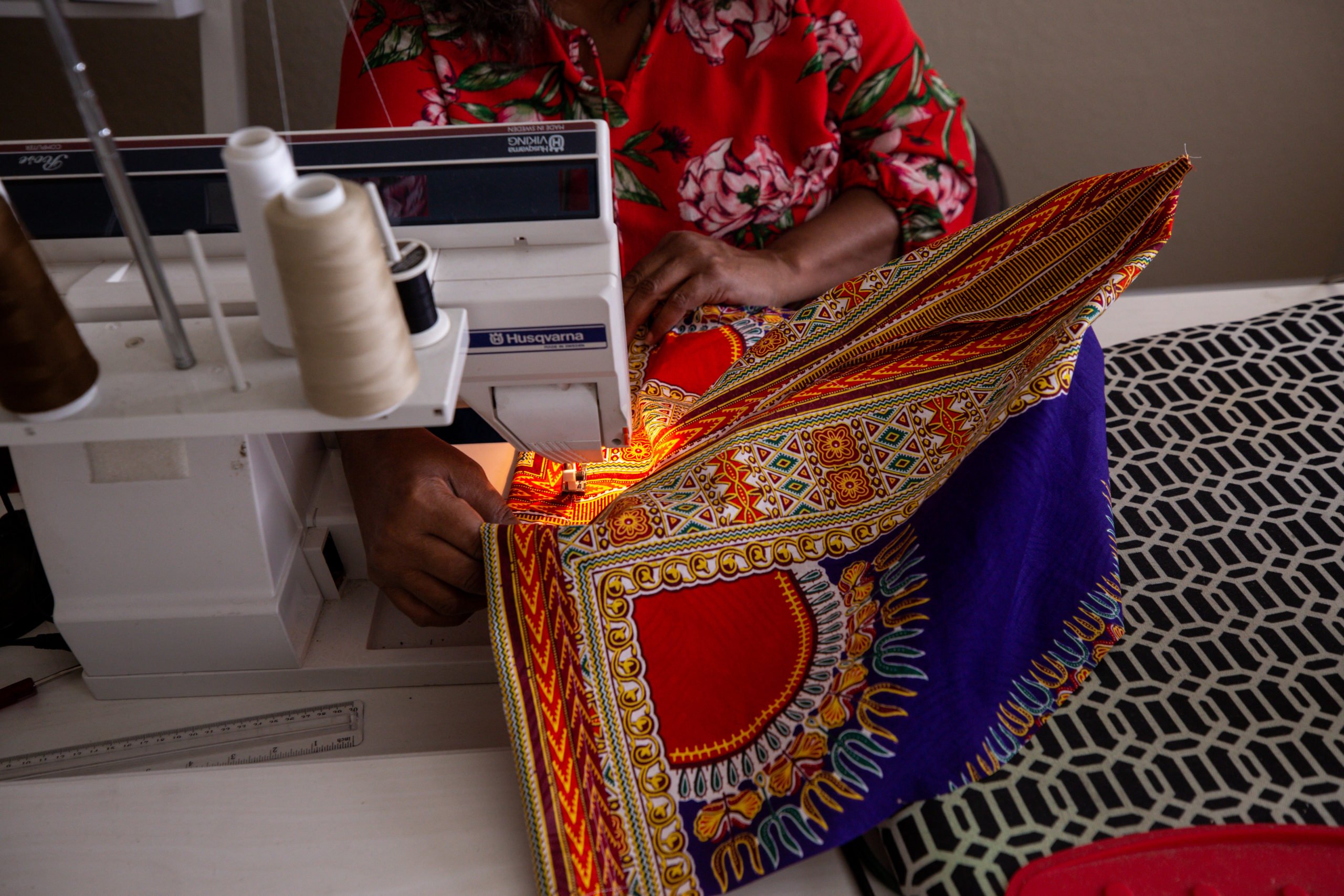 A Black woman, seen as her torso and hands working on a sewing machine, with colorful red and gold fabric being fed through.