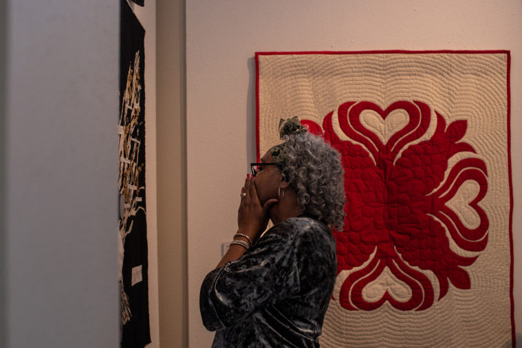 A black woman with curly white and gray hair contemplates quilts hanging on the wall, wearing a thoughtful expression.