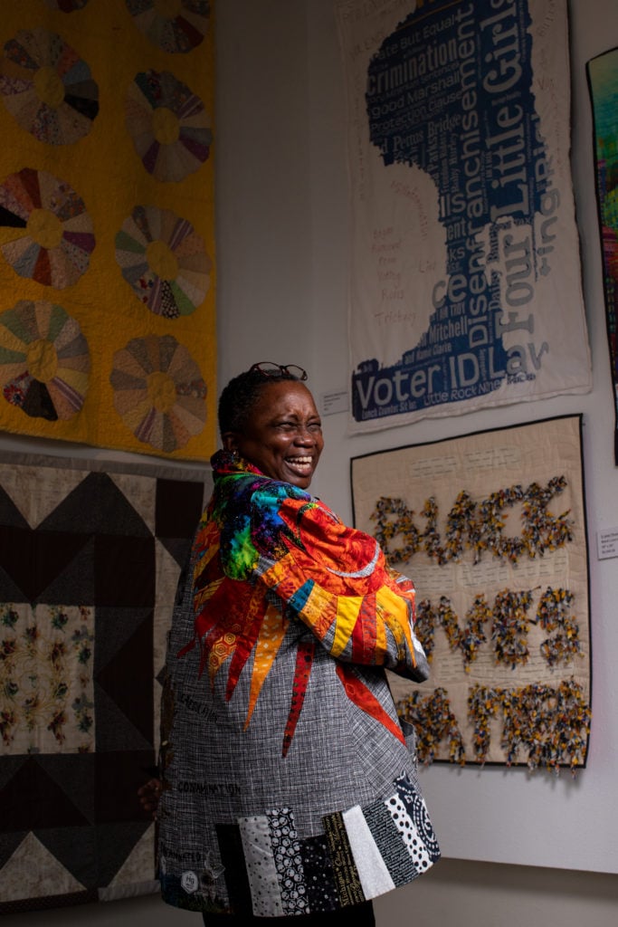 A black woman smiles, wearing a unique colorful quilted jacket. Quilts are hanging on the wall around her.
