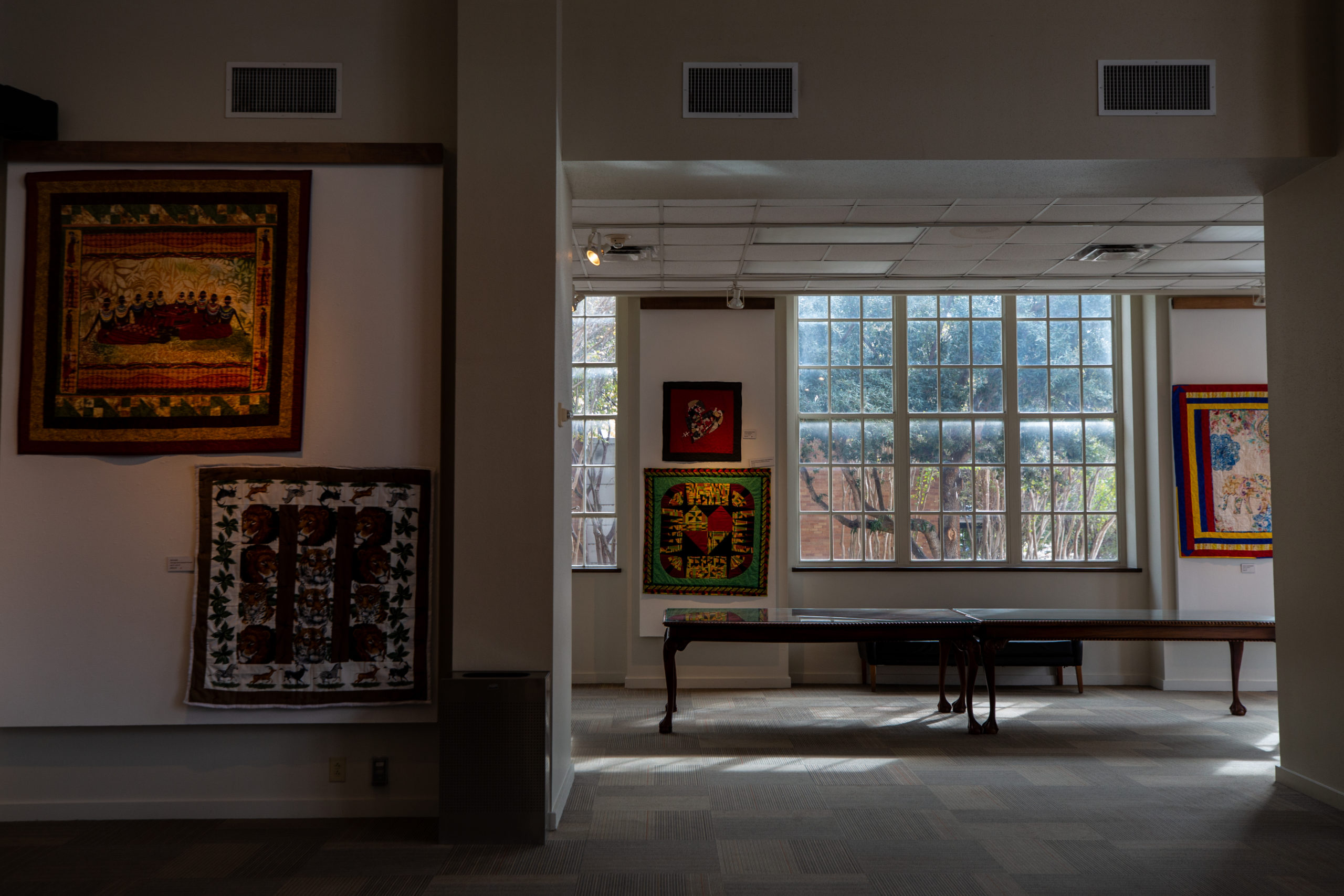 Colorful, artistic quilts hang on the walls of an airy, open gallery with large windows. 