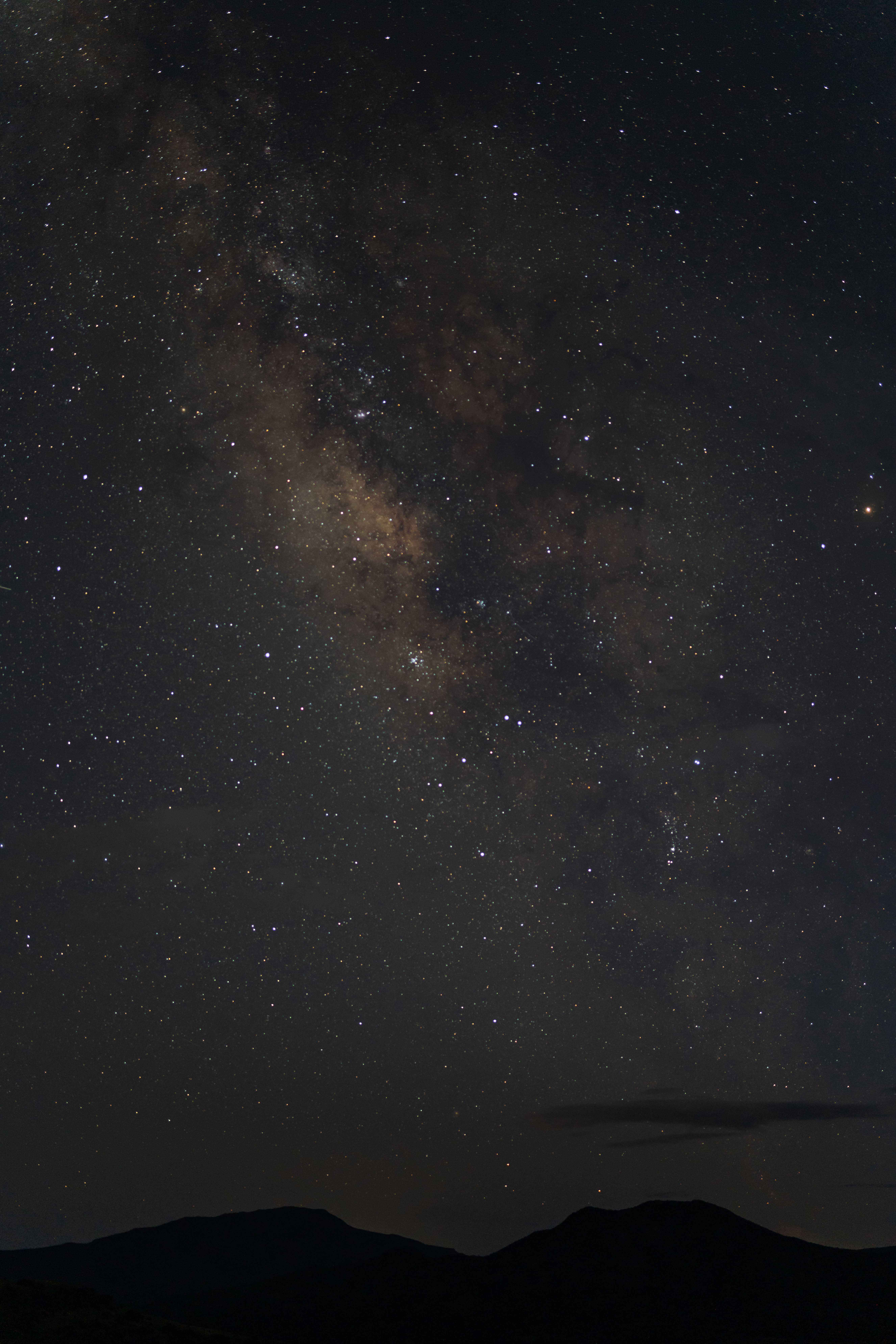 The starfield is deep and dense with milky pools of light speckling the sky. Silhouettes of the mountains can be seen at the horizon.