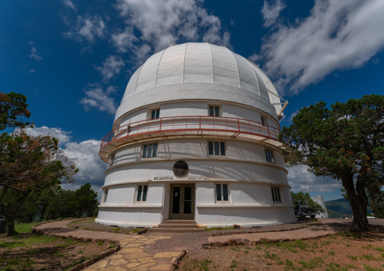 A multi-story white telescopic observation dome stands under a big partly cloudy sky, set among Texas scrub.