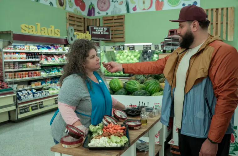 A white woman with curly hair looks on suspiciously as Mo Amer, a Palestinian man, offers a sample of hummus on a pita chip, in a grocery store TV set.