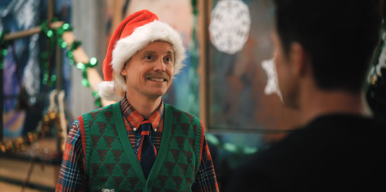 David Hornsby, a white man with a mustache, wears a red and white Santa hat and a v-neck sweater with green and black triangles over a tie. He's smiling at a coworker, seen to the right of him. 