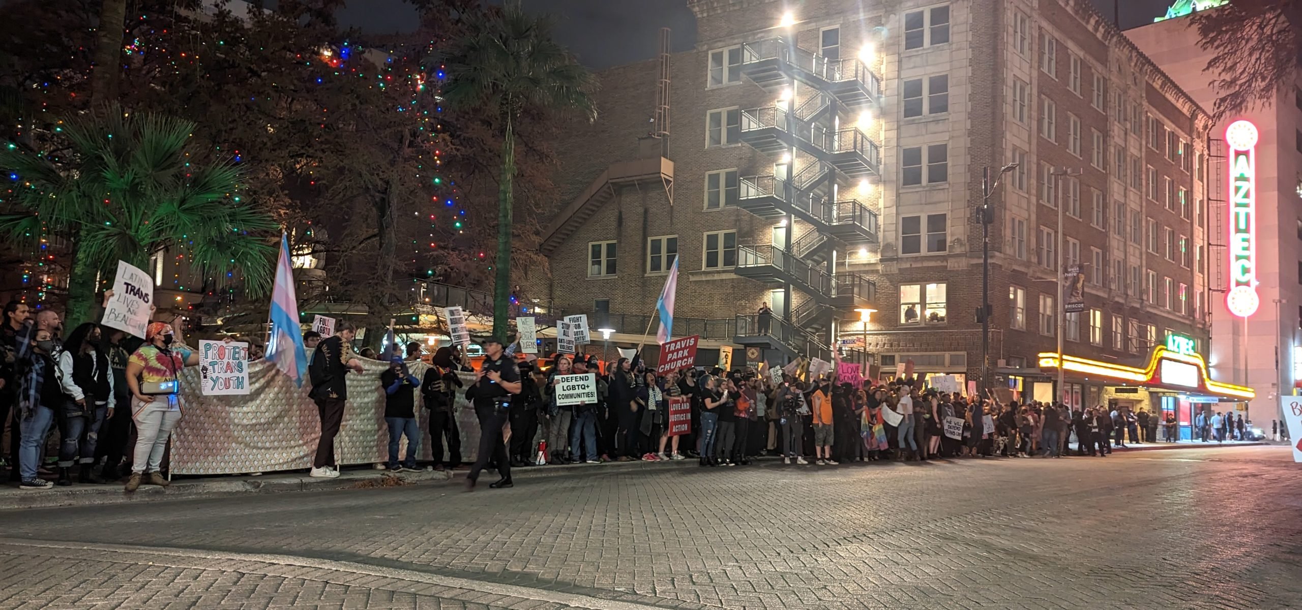 A long row of activists holding signs and trans pride flags at a protest outside a drag event at the Aztec Theater in San Antonio, Texas.