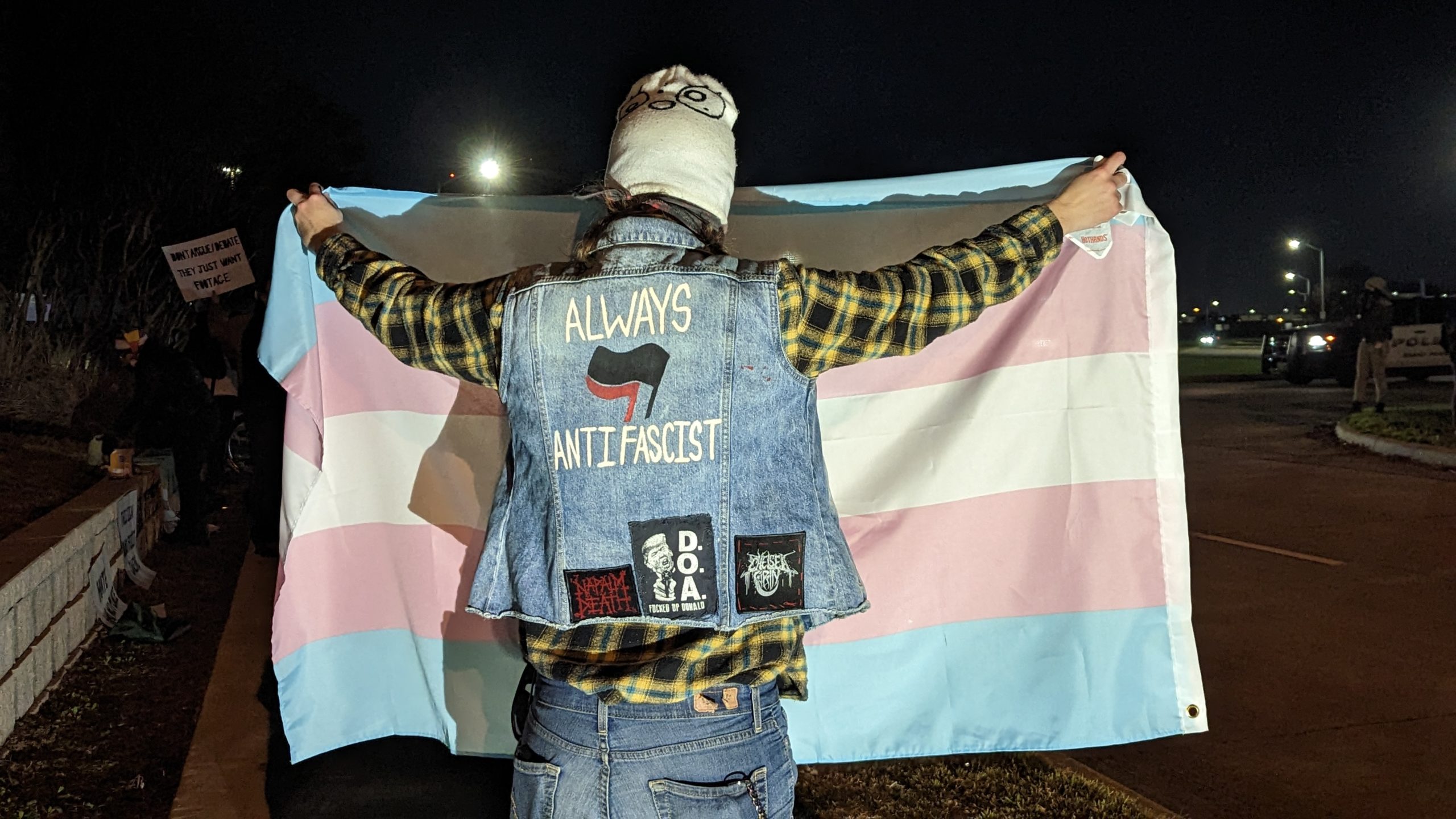At a protest outside a Christmas-themed drag show in Grand Prairie Texas, an activist holds a trans pride flag, wearing a denim jacket decorated with several patches including an Always Antifascist back patch which includes the red and black antifascist flags.