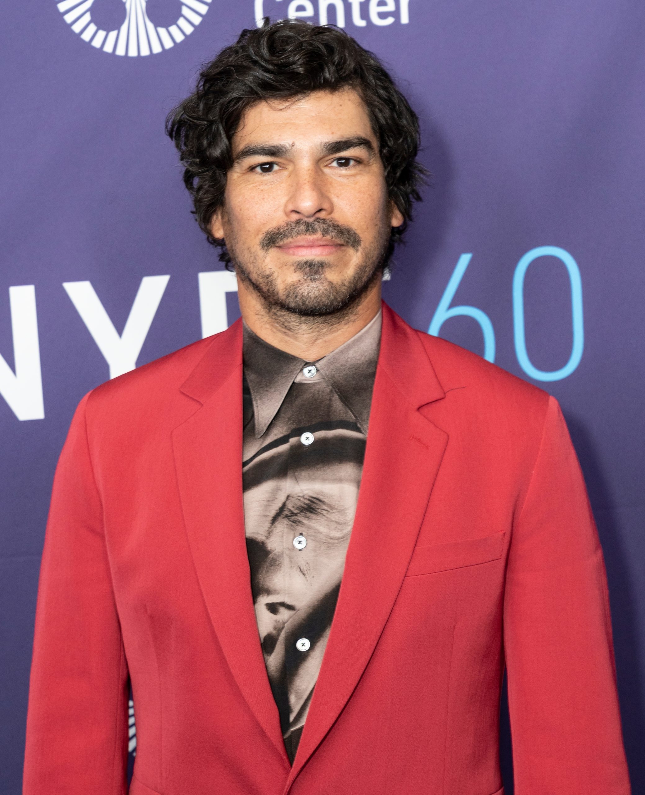Raul Castillo, a Latino man with dark hair, slight beard and mustache, smiles, wearing a red suit jacket over a brown silk shirt.