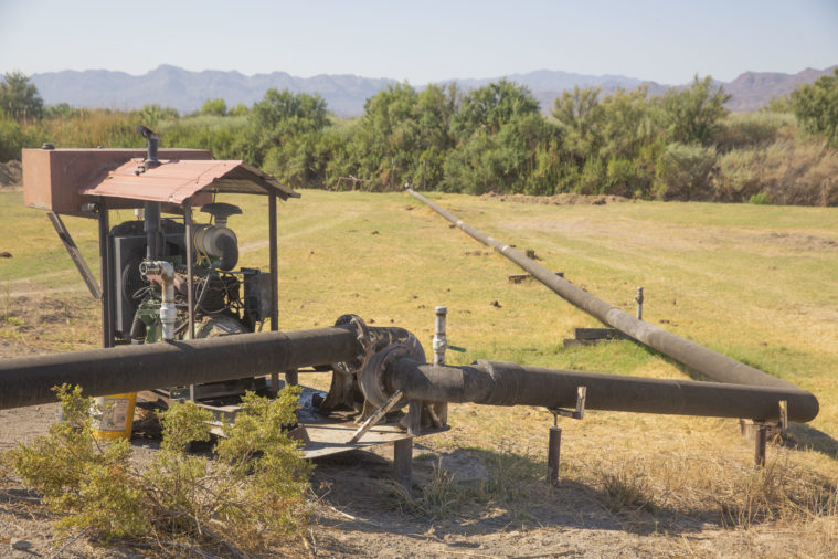 A pump with its own roof, attached to irrigation piping, sits in green field in a lush valley.