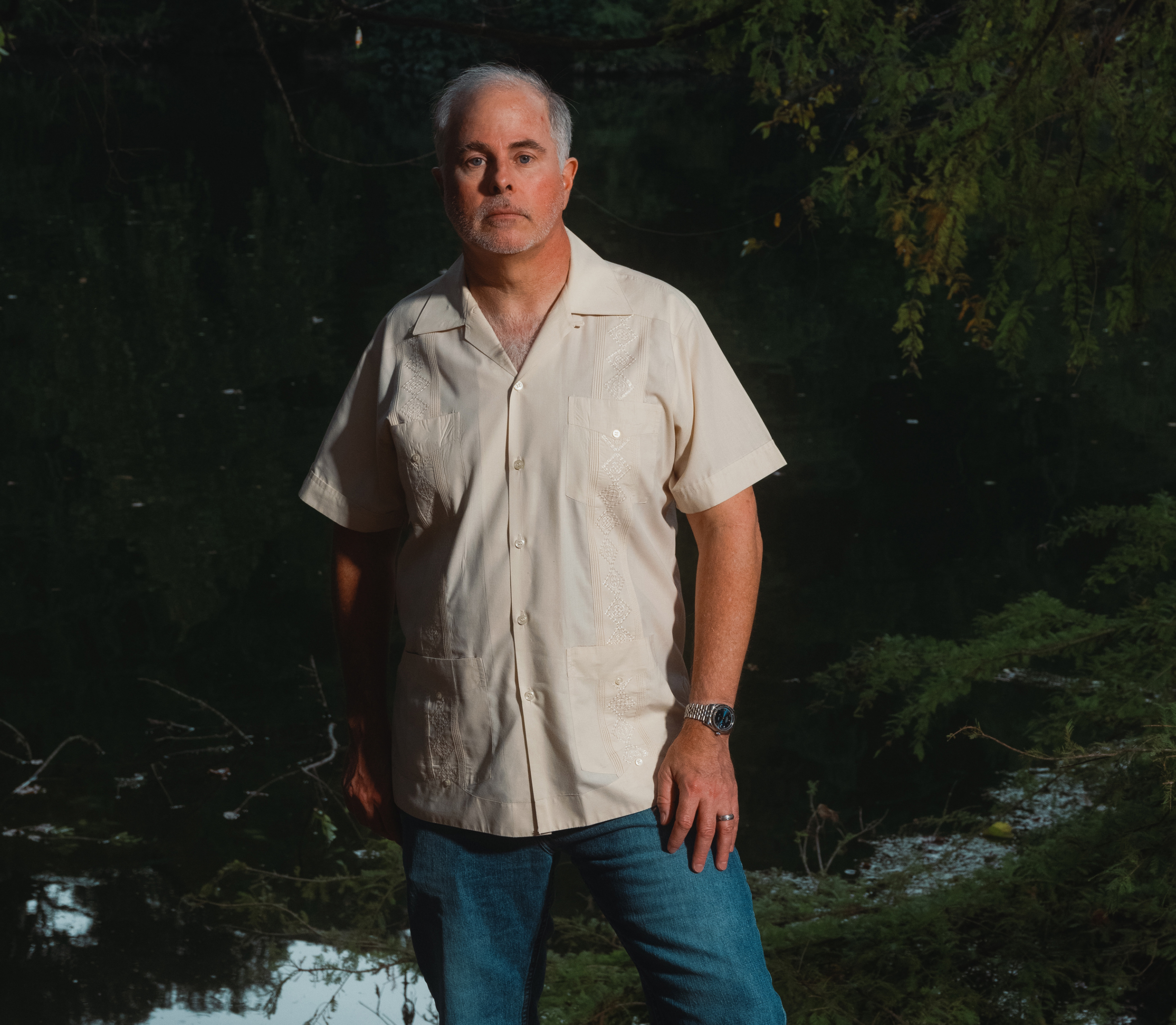 Todd Voteller, wearing a short-sleeve white button-down and blue jeans, stands in front of a placid river dotted with green plants.