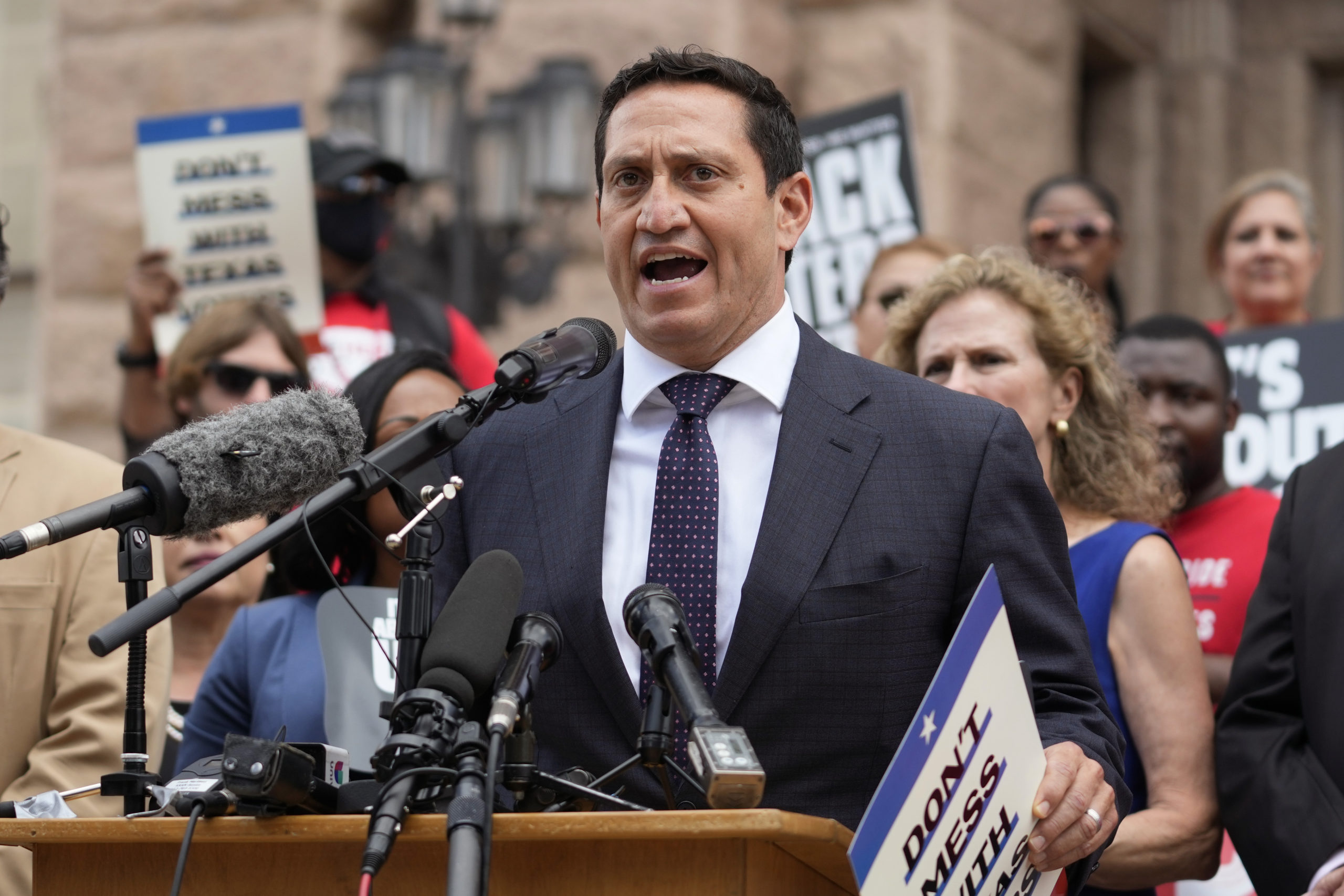 Wearing a blue suit and tie, Trey Martinez Fischer speaks at a podium, surrounded by a diverse crowd of supportive activists.