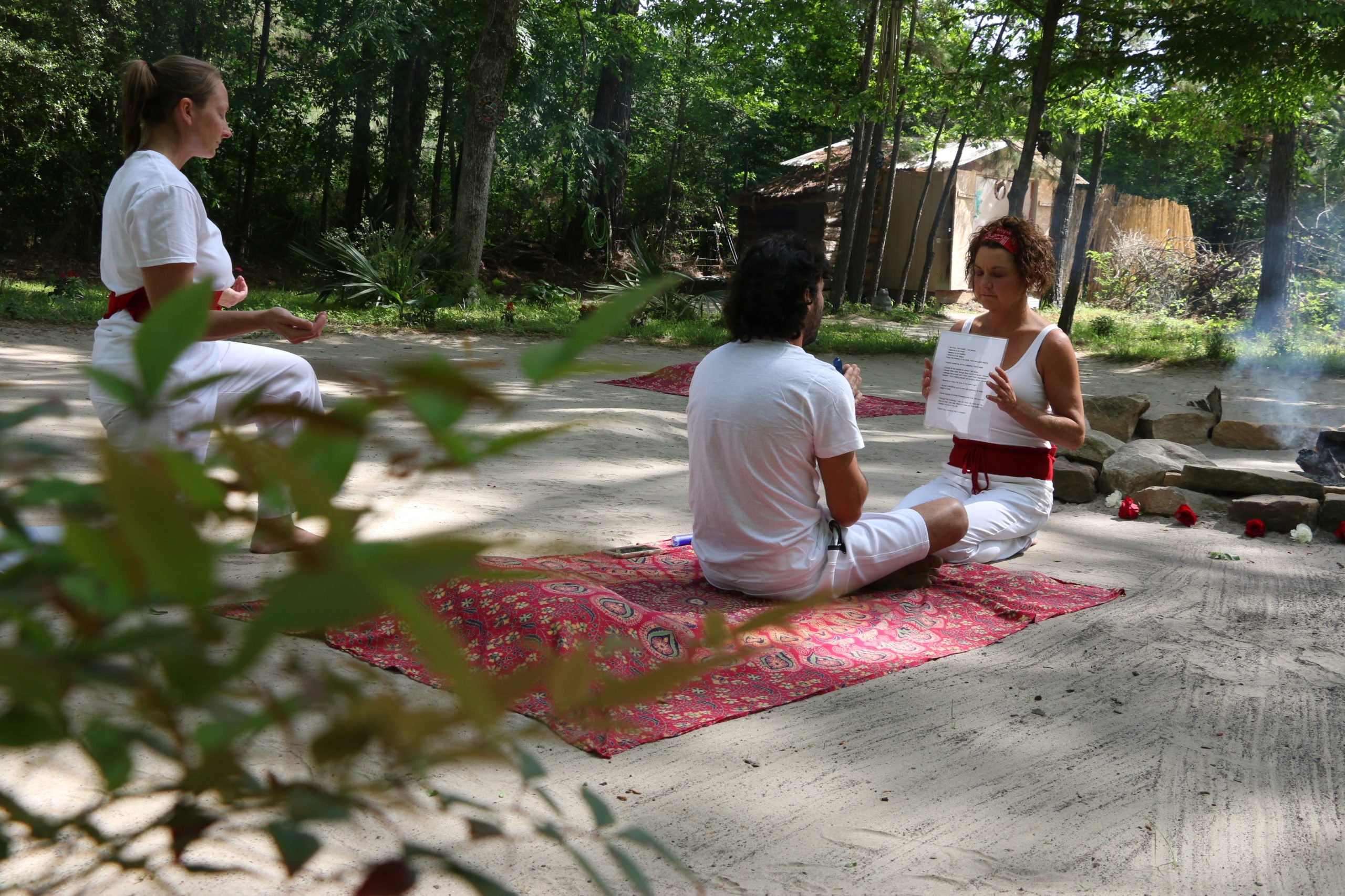 Kneeling, Brooke Tarrer, a white woman woman in white clothes with a red sash, holds a printout with a prayer on it, in front of a person seated cross-legged on a rug, about to smoke bufo. A smoldering fire pit is nearby, and another congregant crouches watching behind them.