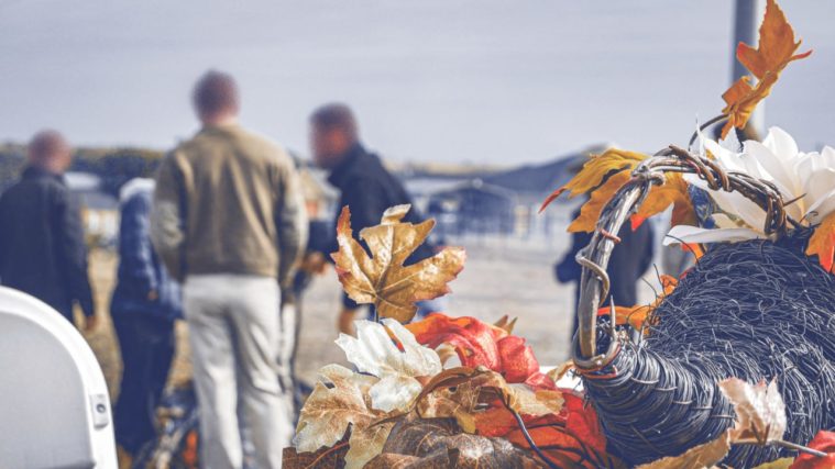 A photo shows a cornucopia and other Thanksgiving decorations while fascsts with blurred faces gather behind it. In the far background is a car port and fence of a neighboring property to where Jason Lee Van Dyke hosted Patriot Front.