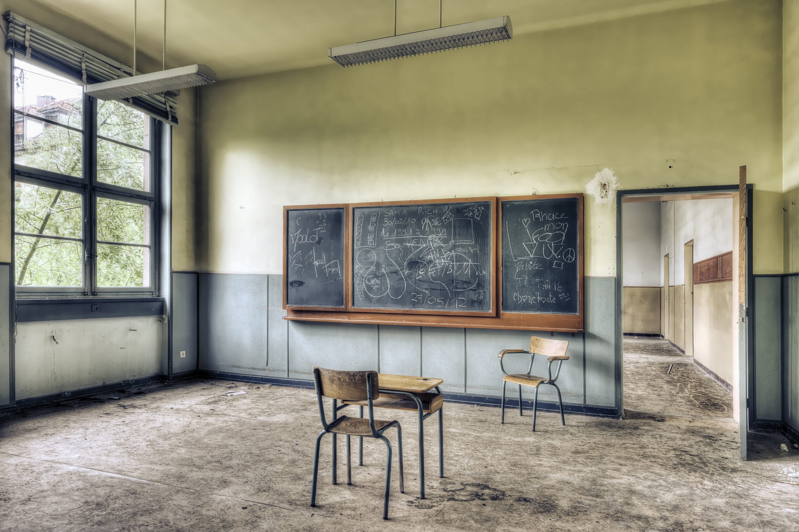 A dusty abandoned classroom, with a worn out looking blackboard, one battered desk-chair and another single chair. A doorway leads into another abandoned room.